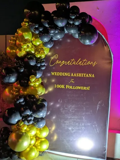 I design branded backdrops for corporate milestones, like this one for a company celebrating 100k followers. The design includes a custom printed backdrop with the company's message, framed by a stylish black and gold organic balloon garland.