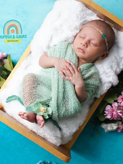 A newborn girl sleeps peacefully in a miniature bed, dressed in a soft mint green outfit. The surrounding blue and pink flowers add a gentle pop of color to the scene.