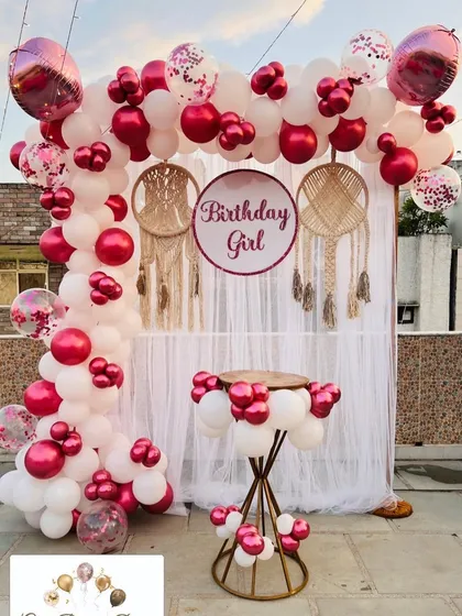 A stylish birthday photo booth on a terrace, with a white curtain backdrop, macrame details, and a vibrant pink and white balloon arch.