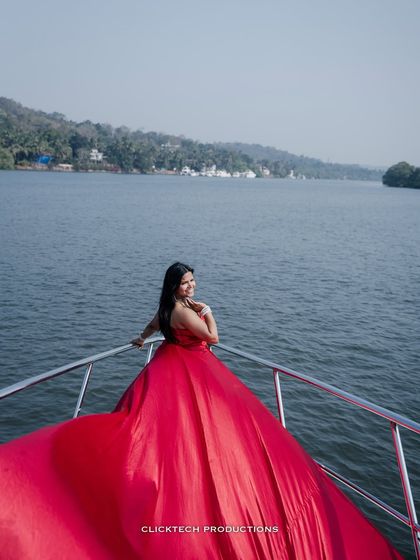 The bride-to-be looks back with a smile from the bow of a yacht, her red gown creating a stunning visual against the water and shoreline.