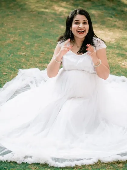 A fun and expressive moment from a park photoshoot. The mother-to-be, seated on the grass in a beautiful white gown, shares a joyful, candid laugh.