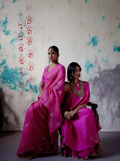 Two models in vibrant pink, showcasing different styles. One wears a delicate organza saree, while the other is in a solid silk ensemble with a heavily embroidered neckline.