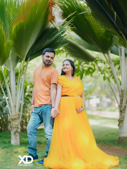 A happy couple posing back-to-back amidst lush palm trees, with the mother-to-be in a vibrant yellow gown.