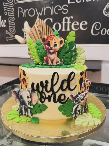 A "Wild One" themed cake perfect for a first birthday. This jungle cake features a baby lion cub surrounded by other safari animals and tropical leaves.