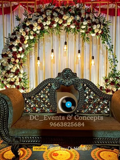 A beautiful engagement stage setup with a floral arch of red and white roses, complemented by an ornate traditional sofa and hanging industrial-style bulbs.