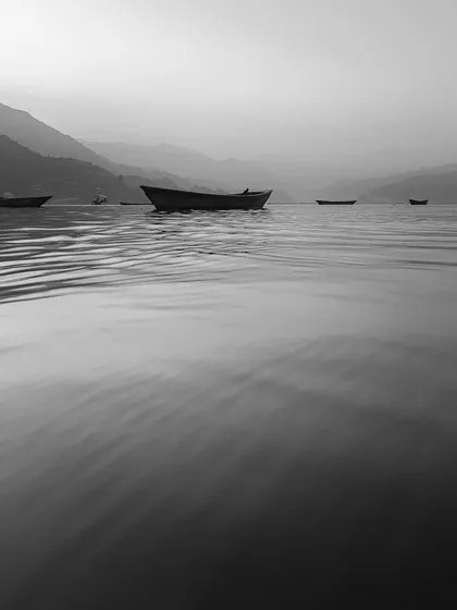 A few boats rest on calm, rippling water under a hazy sky. This minimalist and serene black and white photograph focuses on the subtle textures of the water and the quiet atmosphere.