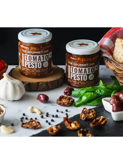Sundried Tomato Pesto jars by The Gourmet Jar, surrounded by ingredients like tomatoes, olives, and focaccia bread. This composition helps customers imagine how they can use the product.