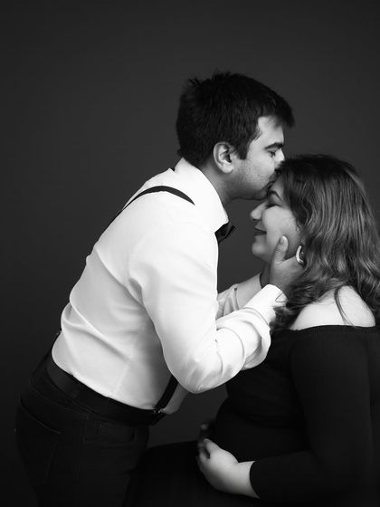 A timeless black and white version of the same moment, where the husband gently kisses his wife's forehead. The monochrome tones emphasize the intimacy and emotion of the portrait.