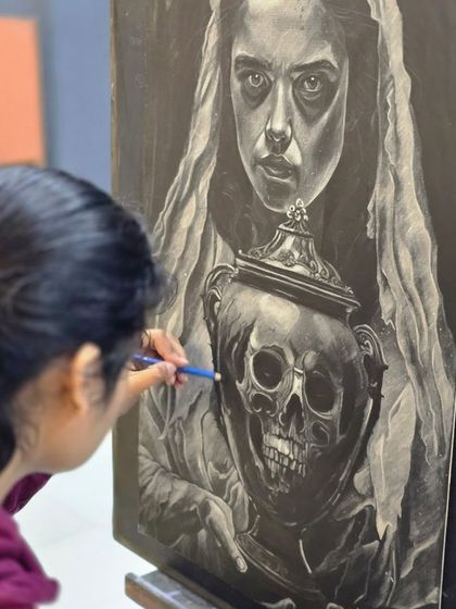 A student works diligently on a complex and moody charcoal portrait, using a pencil to add fine, bright highlights to the skull.
