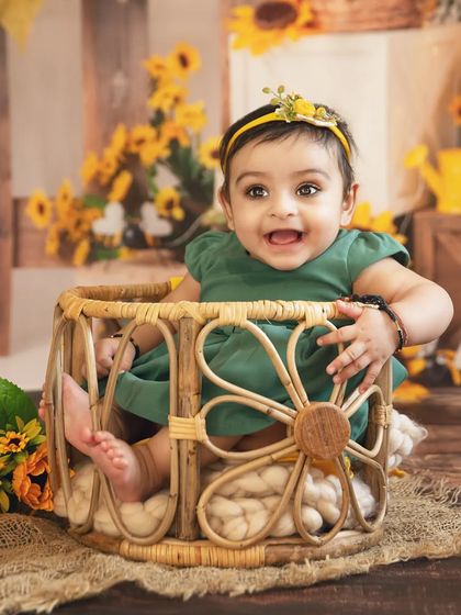 A smiling baby girl in a green dress sits in a wicker basket during her sunflower-themed shoot.