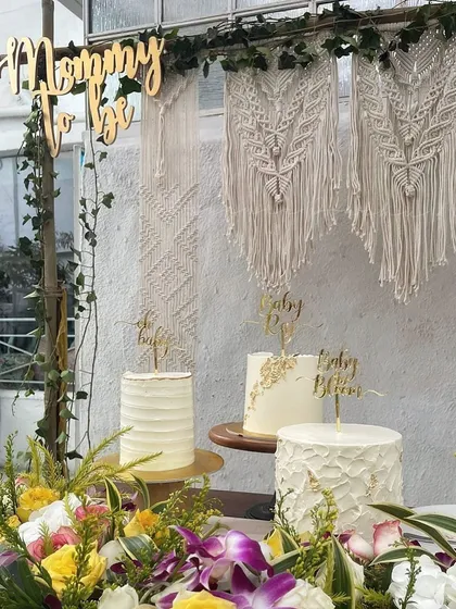 The details of a boho-themed baby shower, featuring a custom macrame art piece and a trio of elegant cakes. The 'Mommy to be' sign adds a lovely, personal touch to the dessert table.