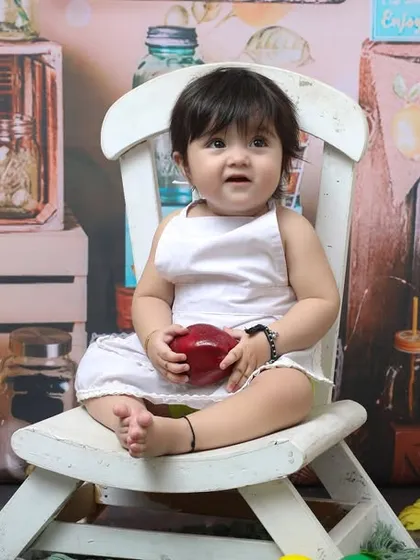 Holding an apple in a charming, rustic setup, this one-year-old is the star of his birthday shoot. The white chair and fruit-stand background create a wholesome and sweet portrait.