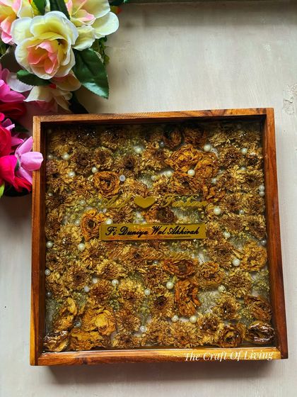 A full view of a 12x12 inch wooden frame filled with preserved yellow marigold flowers from a varmala. The couple's names are elegantly displayed on a golden acrylic strip, adding a touch of luxury.