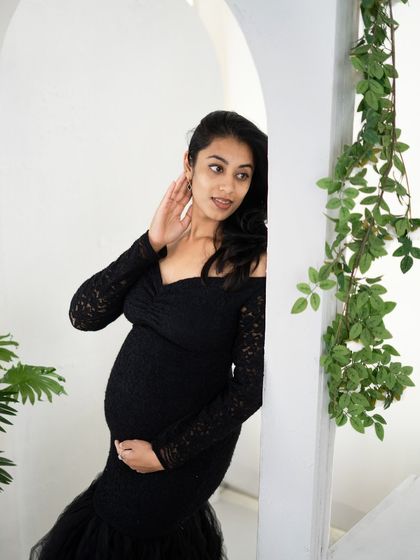 A candid and elegant portrait of a mom-to-be in a black lace gown, peeking out from behind a white archway in our studio.