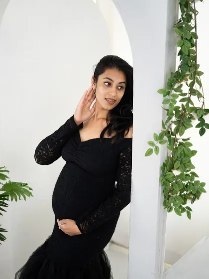 A candid and elegant portrait of a mom-to-be in a black lace gown, peeking out from behind a white archway in our studio.
