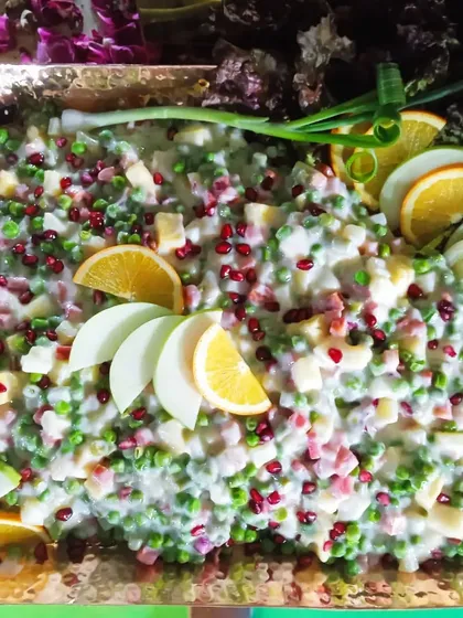 A creamy Russian salad mixed with peas, carrots, and apple, topped with fresh pomegranate seeds and orange slices for a refreshing taste.