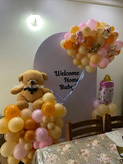 A heartwarming 'Welcome Home Baby' decoration featuring a heart-shaped cutout on the backdrop. The theme is brought to life with teddy bears and balloon arrangements in peach, pink, and orange.