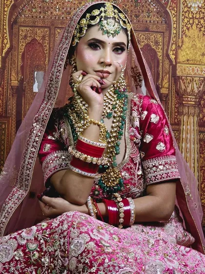 A royal bridal look featuring a pink and red lehenga with heavy embroidery, complemented by a large green-beaded Kundan jewellery set.