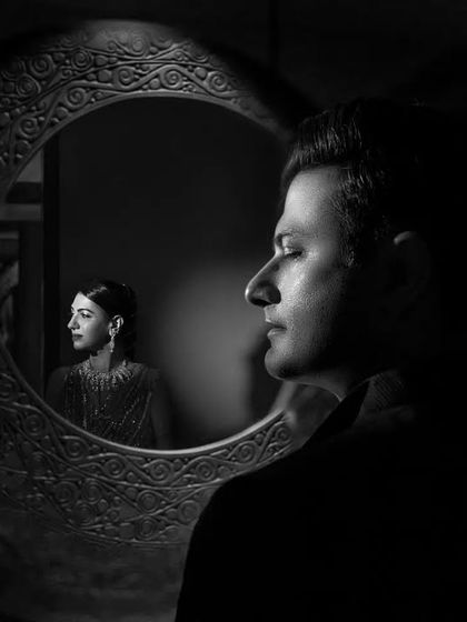 A powerful black and white portrait using a mirror for reflection. The groom is in the foreground with his eyes closed, while the bride's reflection is captured in the mirror, creating a thoughtful and layered composition.