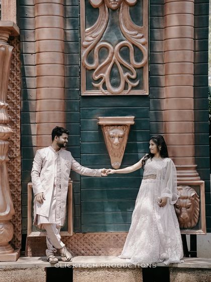 A couple in traditional wear holds hands, posing against a unique wall with large, classical-style carvings.