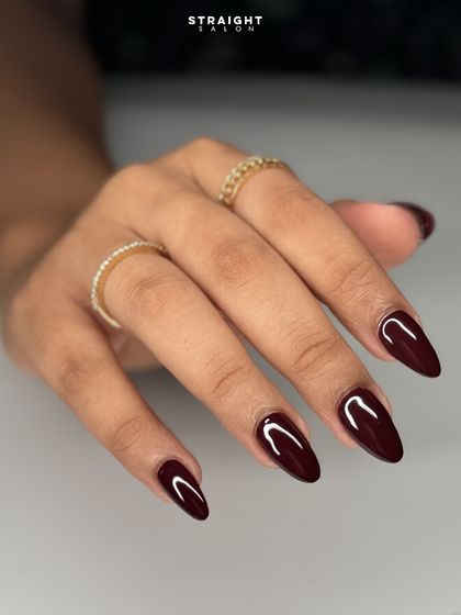 A close-up of the glossy, deep burgundy manicure. The almond shape and flawless finish create a look of pure elegance.