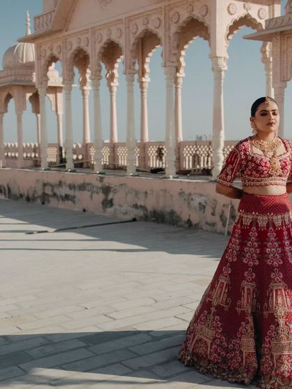 A majestic portrait of the bride in a traditional red lehenga, standing on a palace rooftop with the clear blue sky behind her.