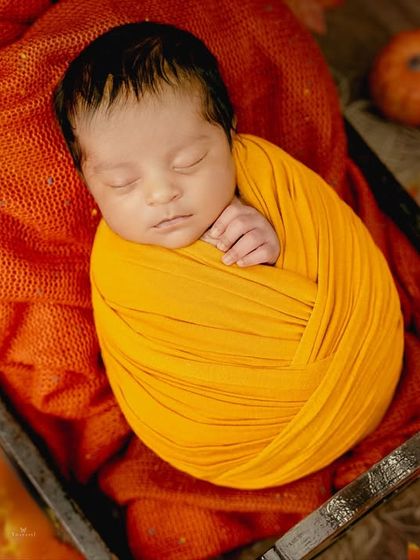 A tightly swaddled newborn in a bright yellow wrap, resting on an orange blanket amidst fall leaves, highlighting the vibrant colors of the season.