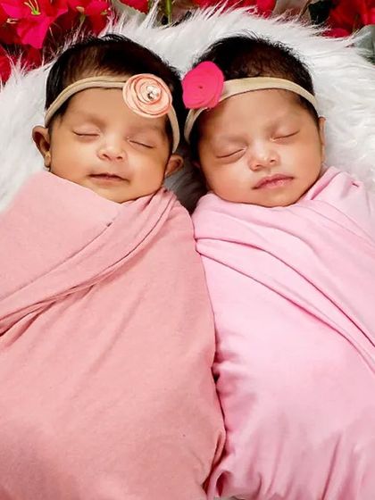 A close-up shot of twin baby girls, swaddled in pink and nestled together among vibrant flowers. Their peaceful expressions show how comfortable they are.