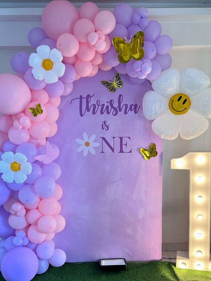 A lovely first birthday setup for Thrisha with a daisy and butterfly theme. The lavender backdrop is framed by a pink and purple balloon arch, accented with large daisy balloons and a light-up number '1'.