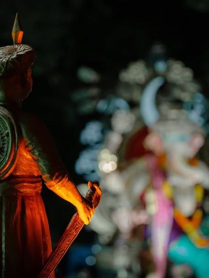 A creative composition focusing on a statue of a Maratha warrior, with the grand Ganesh idol blurred in the background, telling a story of culture and history.