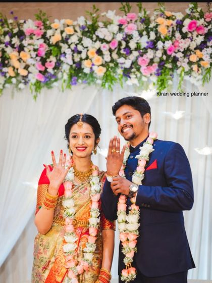 A joyful couple showing off their engagement rings in front of a beautiful pastel floral arch.