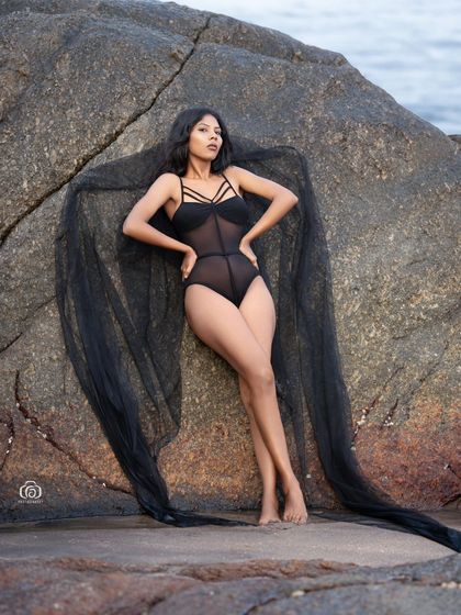 A striking portrait of a model in a black bodysuit, with a sheer black fabric creating wing-like shapes against the rocky background. This is a great example of a creative, high-fashion beach shoot.