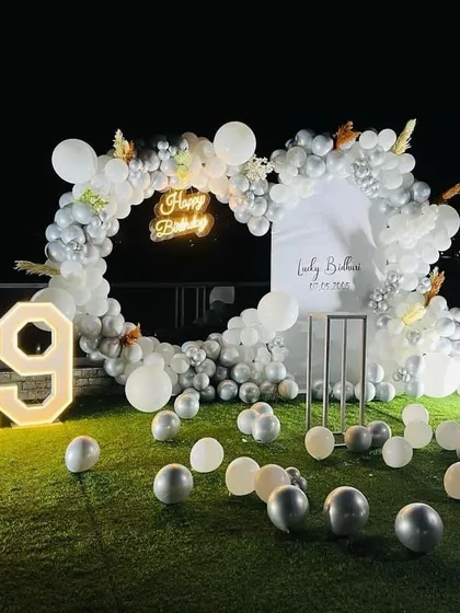 We can take the party outdoors! This elegant 19th birthday setup on a lawn features a white and silver balloon arch, a personalized backdrop, and large marquee numbers, creating a beautiful contrast against the natural greenery.