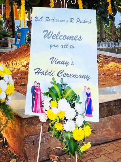 This welcome sign for a haldi ceremony is placed on a stand and decorated with a fresh arrangement of yellow and white flowers, matching the event's theme.