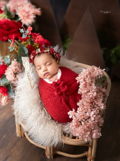 A newborn in a bold red wrap rests in a miniature wicker chair, adorned with a floral headband and surrounded by soft pink and red flowers.