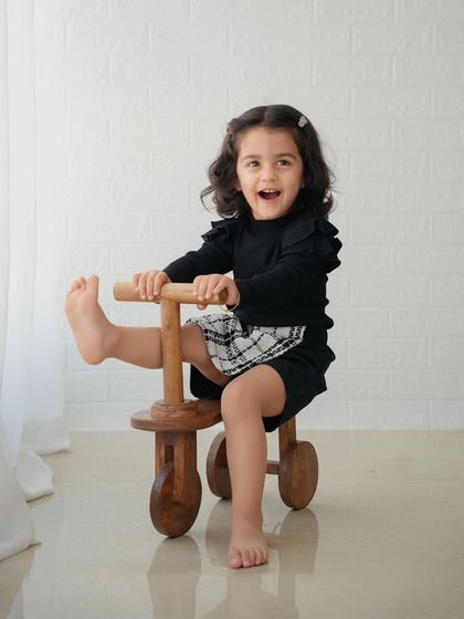 A playful toddler kicks her leg up while riding a wooden tricycle indoors, laughing with excitement.