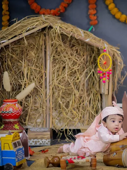 A baby in a pink traditional outfit crawls near a dhol in our rustic Lohri setup, complete with a hay hut and marigold decorations.
