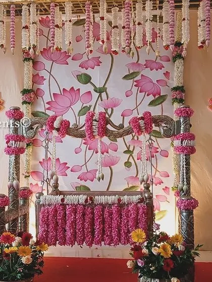 A beautiful naming ceremony design featuring a Pichwai-style lotus painting as the backdrop. The cradle is decorated with pink flowers, and the entire setup is framed by hanging jasmine garlands.