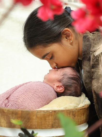A close-up shot of a sister kissing her swaddled newborn sibling in a wooden bucket prop. The red flowers in the foreground add a pop of color and frame the moment beautifully.