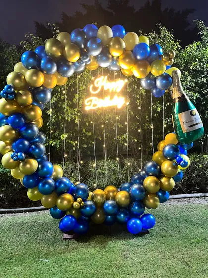 An elegant outdoor birthday celebration setup. The circular frame is decorated with a classy blue and gold balloon garland, complemented by fairy lights and a champagne bottle balloon for a festive touch.