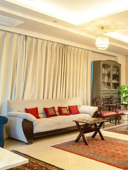 This cozy corner of the living room balances classic and contemporary elements. A neutral sofa is accented with vibrant red cushions, while traditional carved chairs and oriental rugs ground the space in timeless style.