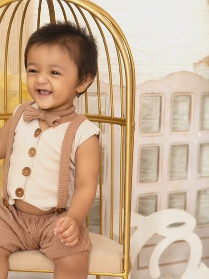 A joyful one-year-old boy laughs while sitting in a golden birdcage chair during his boho-themed session.