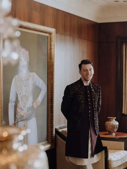 A candid shot of the groom smiling, standing next to a royal painting in a lavishly decorated room.