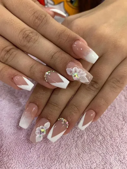A beautiful example of a modern French manicure with 3D floral accents. This design features a soft ombre base, crisp white V-tips, and delicate sculpted flowers with iridescent crystal centers.