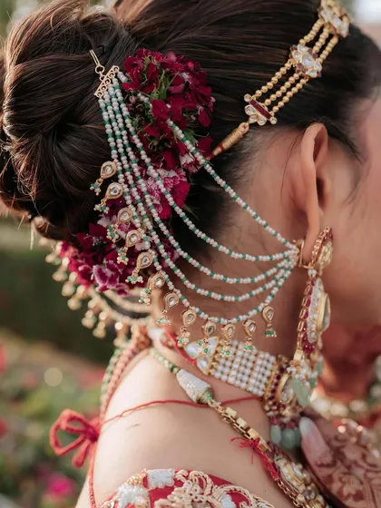 A close up of an intricate bridal bun, adorned with deep red flowers and a multi-layered hair accessory. I focus on creating these detailed updos that not only look stunning but also securely hold the dupatta and jewelry in place.