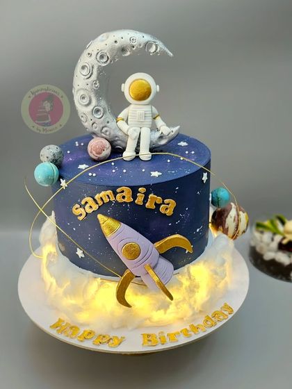 A dreamy galaxy-themed cake with an astronaut sitting on the moon, a rocket, and planets, all illuminated with fairy lights.