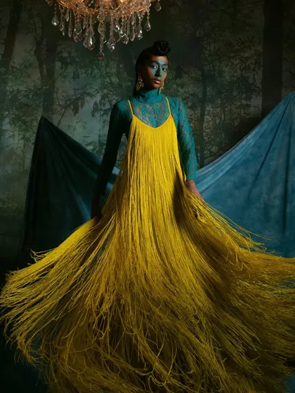 A full-length shot from a conceptual series, featuring a dramatic yellow fringe dress against a forest-themed backdrop. The movement of the fringe is the central focus of this image.