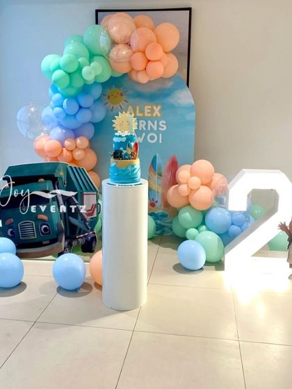 For Alex's second birthday, I put together this unique Trash Truck theme. The setup includes a custom arched backdrop with a beach scene, a cutout of the truck, a light-up number '2', and a pastel balloon garland.