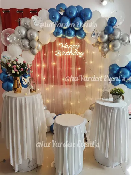 A blue, white, and silver balloon arch framing a set of tables with a fairy light curtain in the background.
