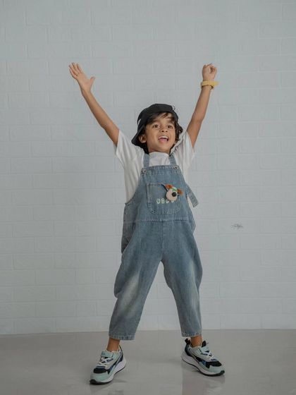 An energetic shot of a boy in overalls and a cap with his arms raised in excitement against a white brick wall.
